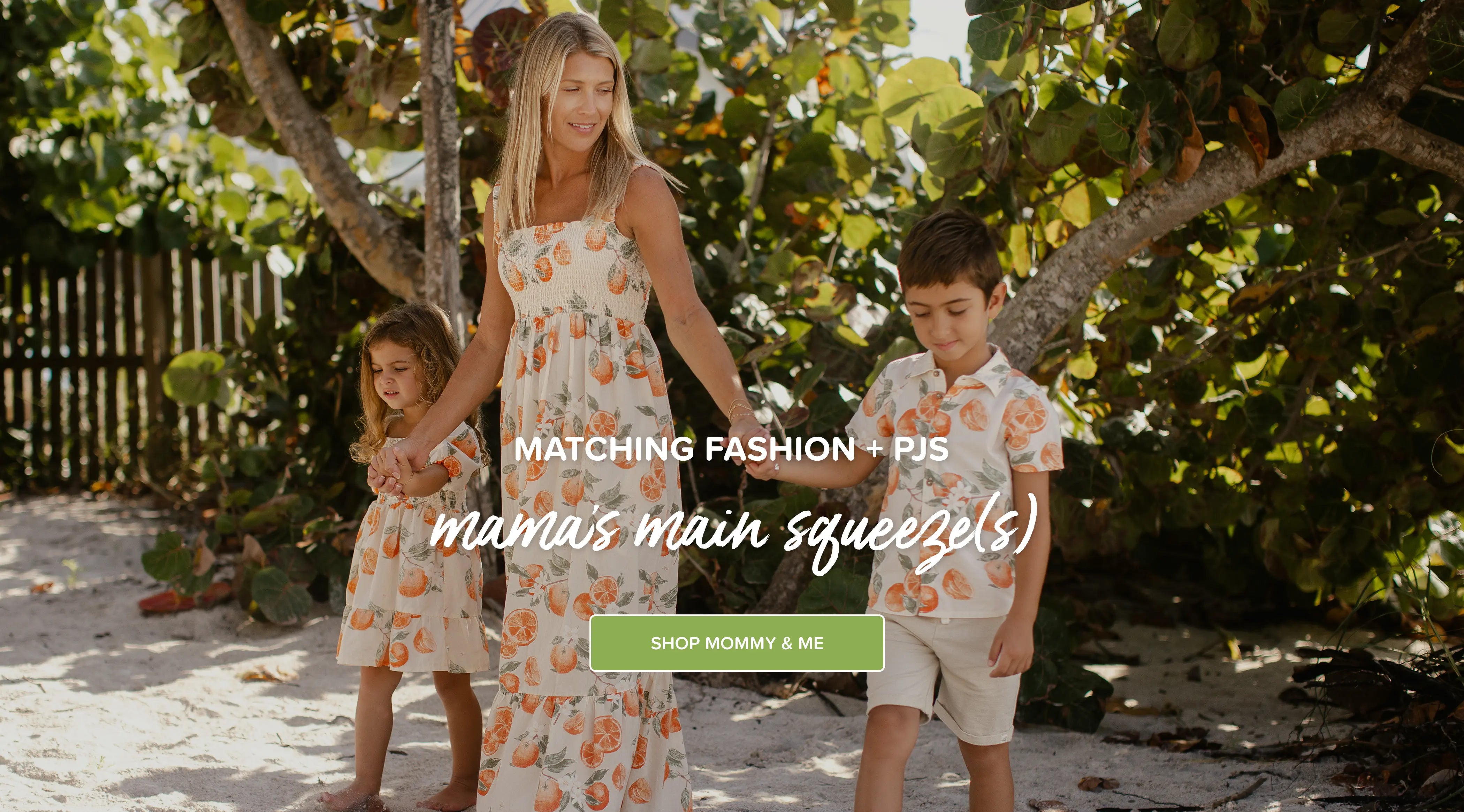 Family of three in matching outfits standing under a tree on a beach. outfits shown include dubrovnik oranges matching girl dress, dubrovnik oranges matching button down shirt & shorts set and dubrovnik matching women's dress