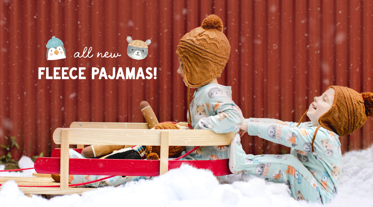 Two children wearing matching light blue patterned fleece pajamas and brown knit pom-pom hats are playing in the snow in front of a red wooden fence. One child is sitting on a wooden sled while the other pushes it.