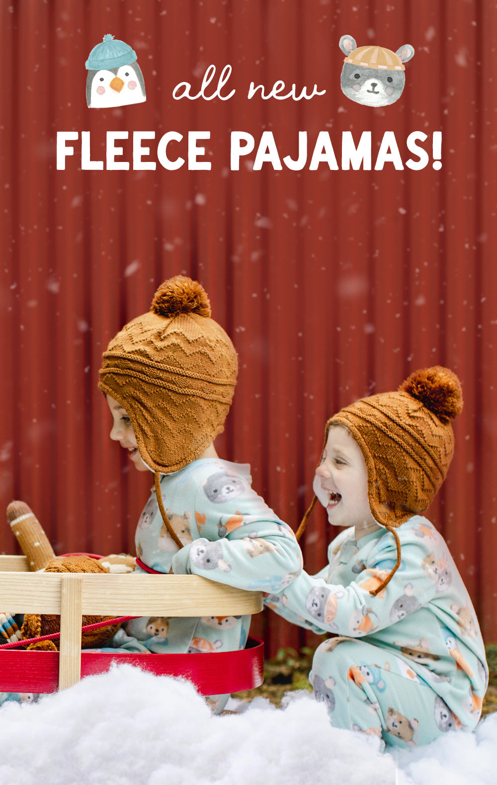 Two children wearing light blue patterned fleece pajamas and brown knit pom-pom beanies sit in a red sled in the snow in front of a red corrugated metal wall. White text at the top reads 'all new FLEECE PAJAMAS!' with penguin and bear illustrations.