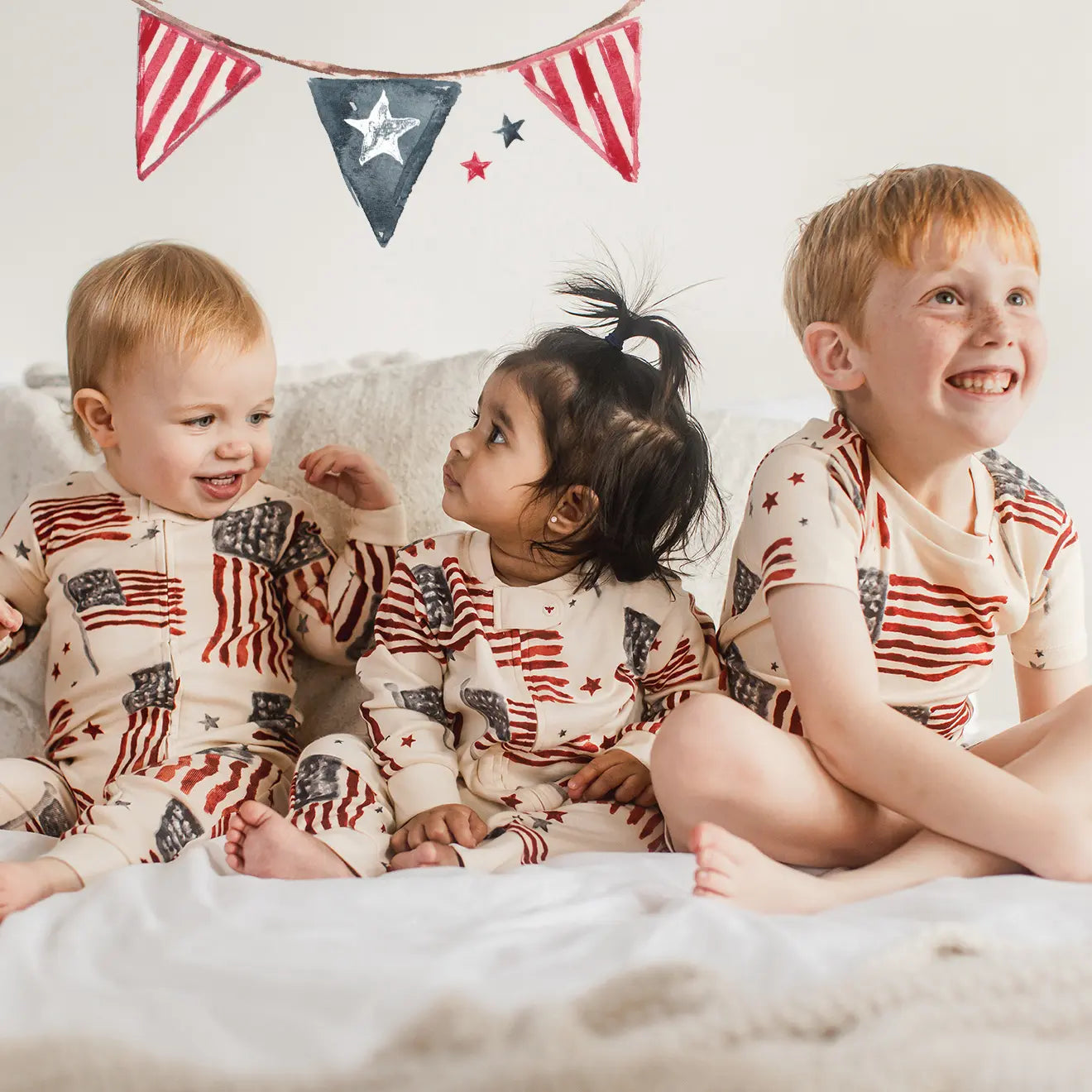 Three children in matching pajamas with American flag design pajamas sitting on a bed.