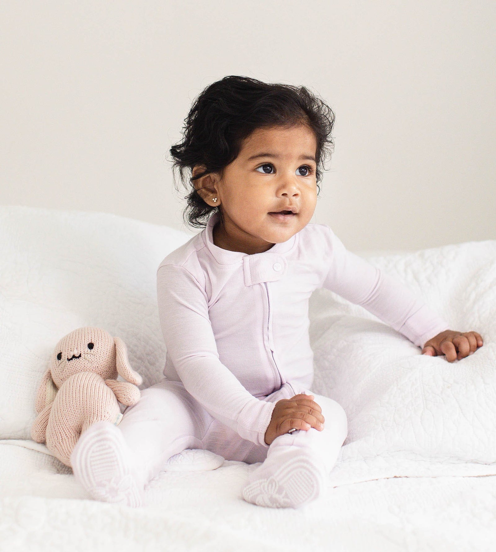 Baby leaning on pillows in a bed wearing one-piece snug fit purple pajamas next to stuffed rabbit