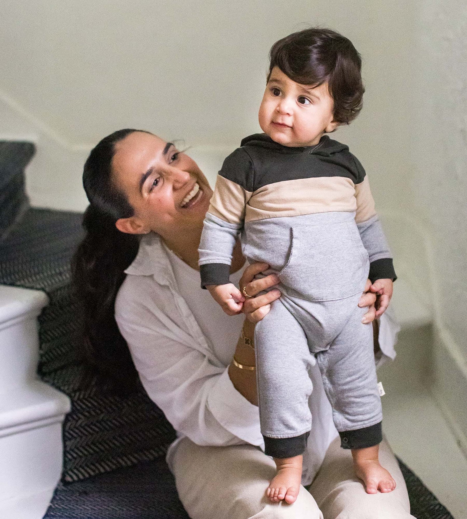 Baby boy standing on smiling mother's lap wearing organic color blocked jumpsuit
