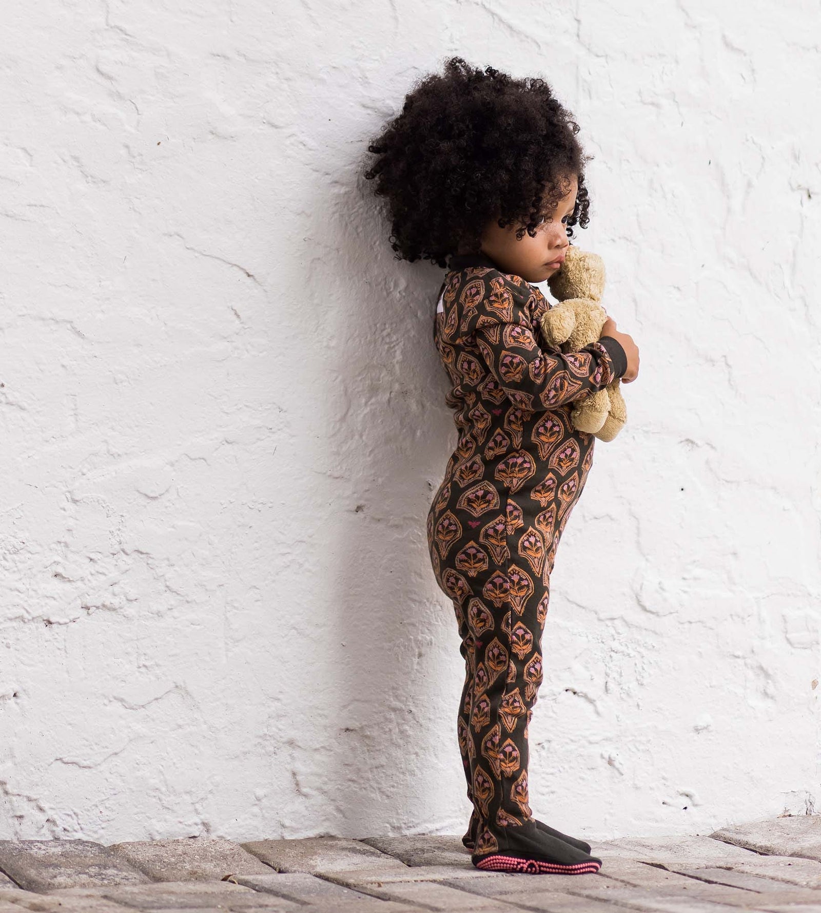 Toddler girl standing near stucco wall holding stuffed bear doll wearing floral arches print one-piece pajamas