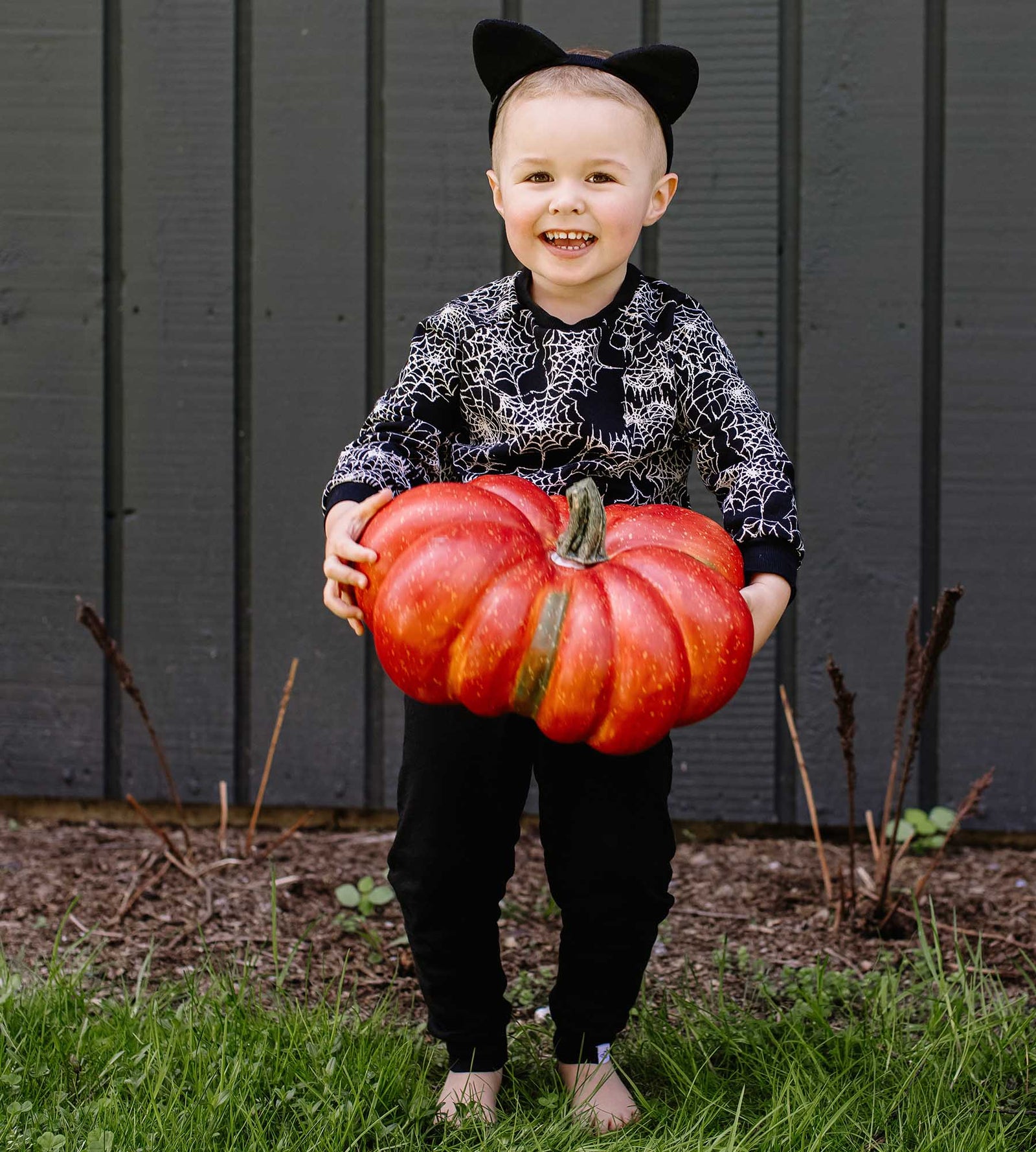 Toddler boy smiling holding an orange pumpkin wearing spiderwebs sweatshirt from Burt's Bees Baby