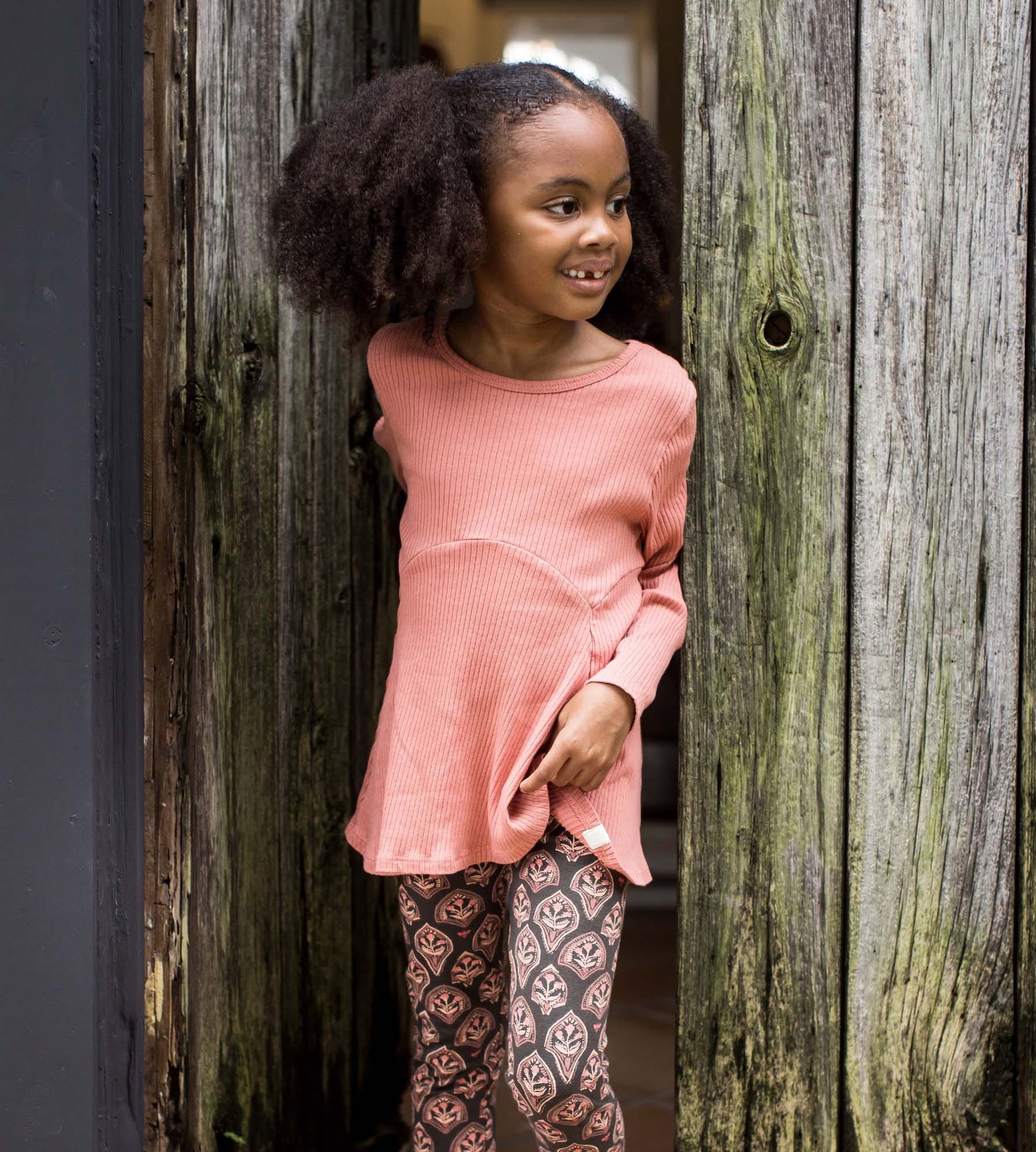 Toddler girl standing by a fence wearing pink ribbed top and floral arches print pants