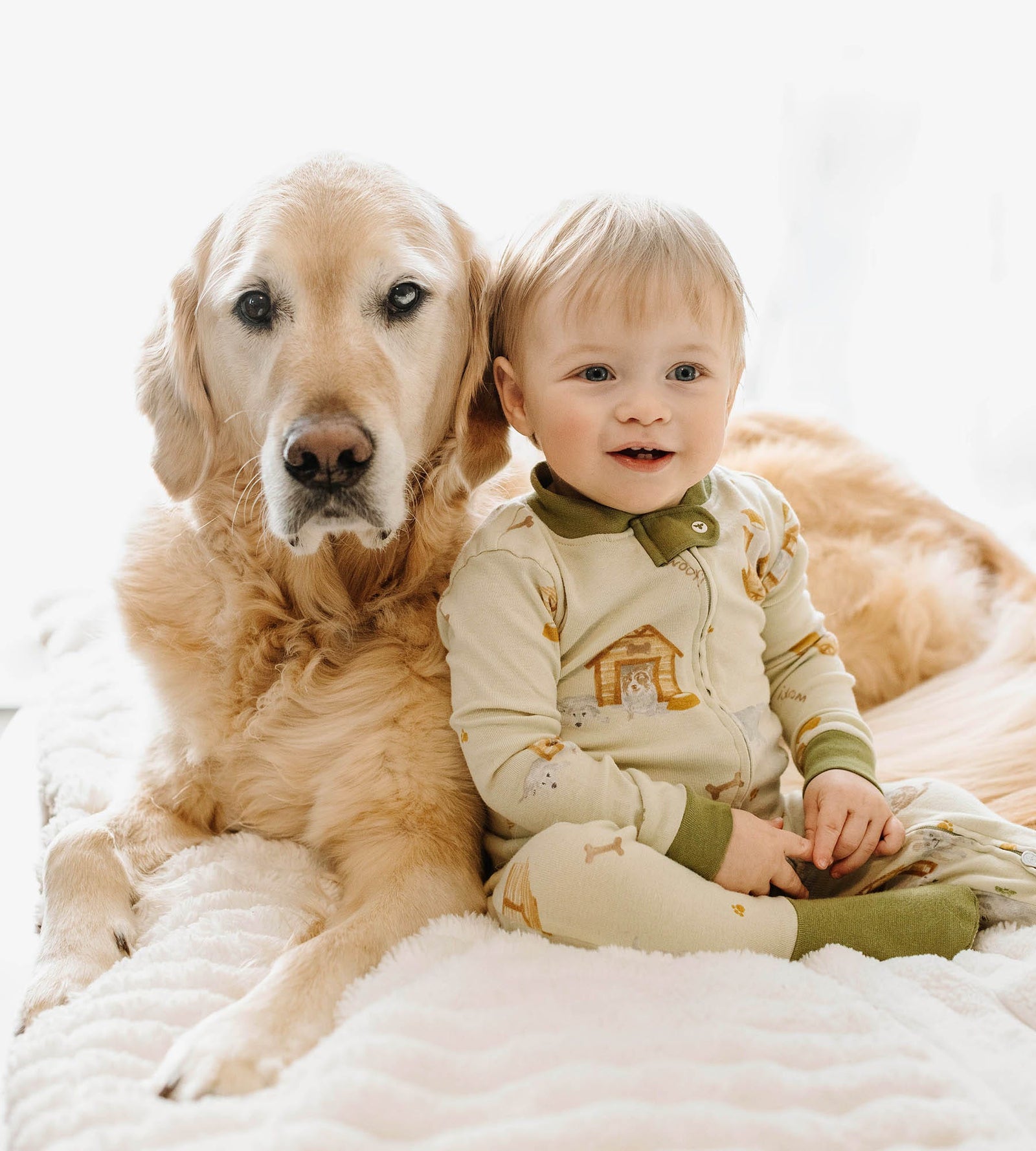 Baby boy wearing dog house print Burt's Bees Baby pajamas sitting on a bed with an old golden retriever