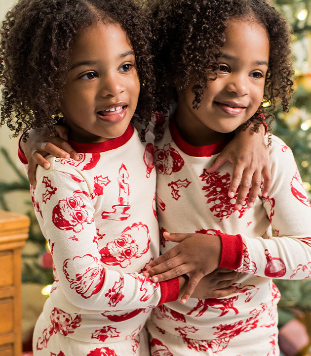 Two young girls smile and hug while wearing matching cream pajamas with red holiday prints.