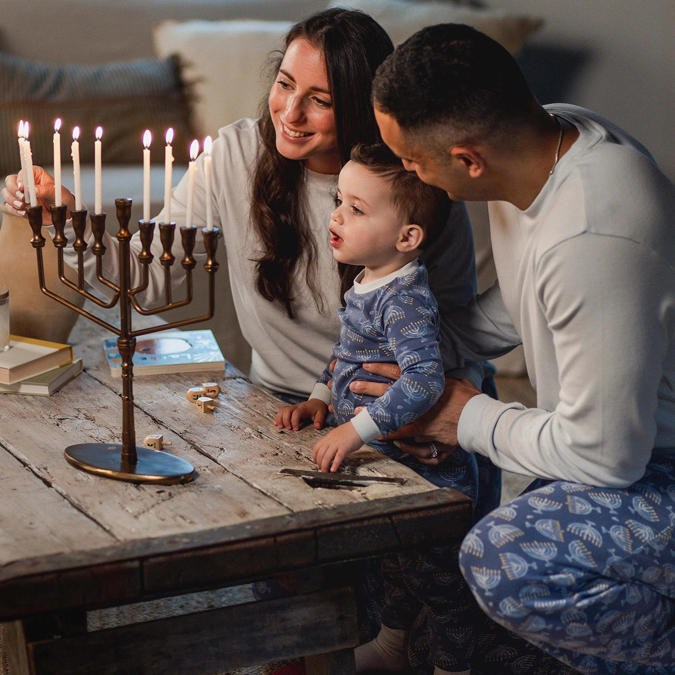 Family of three, including a young child, gathered around a menorah on a wooden table.
