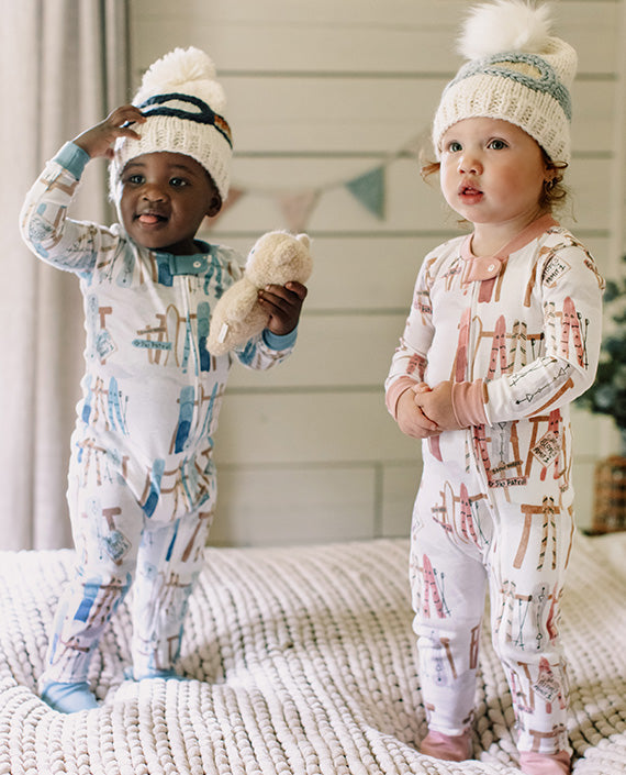 Two children wearing matching sunrise slopes  pajamas and winter hats on a bed.